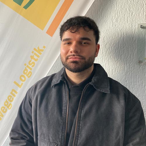 Mann mit dunkler Jacke vor Banner, neutraler Ausdruck, kurze Haare und Bart.