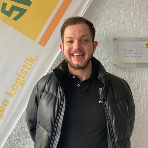 Mann lächelt fröhlich, schwarze Jacke, schwarzes Polohemd, weiße Flagge im Hintergrund.