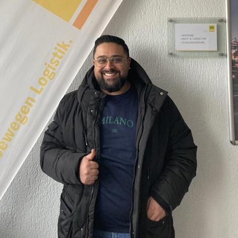 Mann lächelt, trägt schwarze Jacke vor weißer Wand. Schild und Banner im Hintergrund.