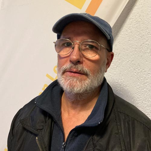 Mann mit Bart und Brille, blauem Basecap; ernster Ausdruck, schwarze Jacke vor heller Wand.