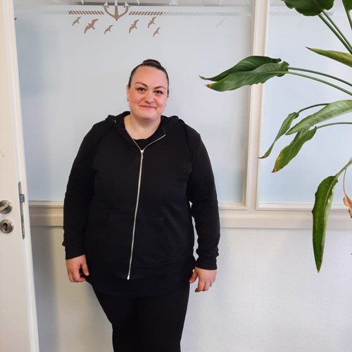 Eine Frau mit dunklen Haaren und schwarzem Hoodie lächelt freundlich in einem hellen Raum