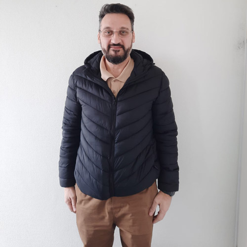 Mann mit Bart und Brille lächelt leicht in brauner Hose und schwarzer Steppjacke