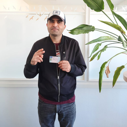 Ein lächelnder Mann mit schwarzer Jacke und Basecap hält einen Ausweis in der Hand