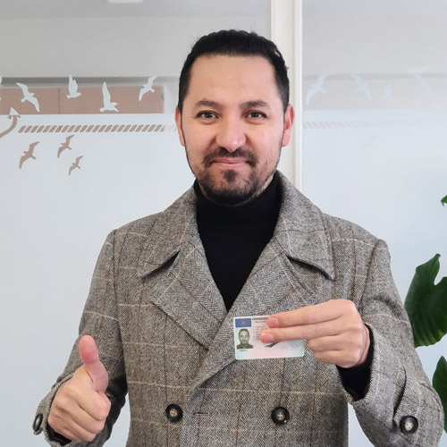 Mann mit dunklen Haaren und Bart lächelt, hält Ausweis und zeigt Daumen hoch