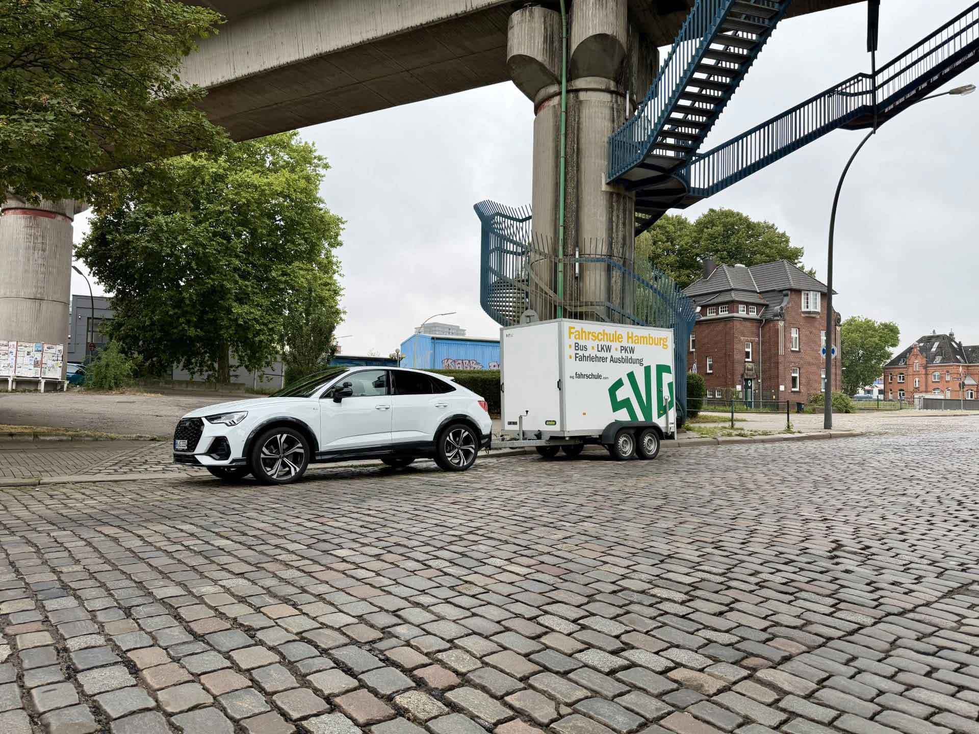 Weisser Audi mit Fahrschulanhaenger auf Kopfsteinpflaster unter einer Bruecke