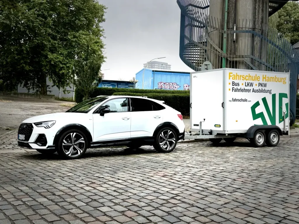 Weisser Audi SUV mit Fahrschulanhaenger der SVG auf gepflastertem Platz